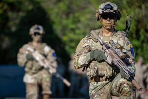 Soldiers in tactical gear holding rifles during a military training exercise in a wooded area.
