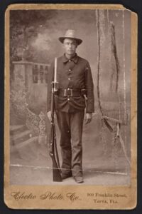 Soldier in historical uniform holding rifle, vintage military portrait, black-and-white photo from early 20th century.